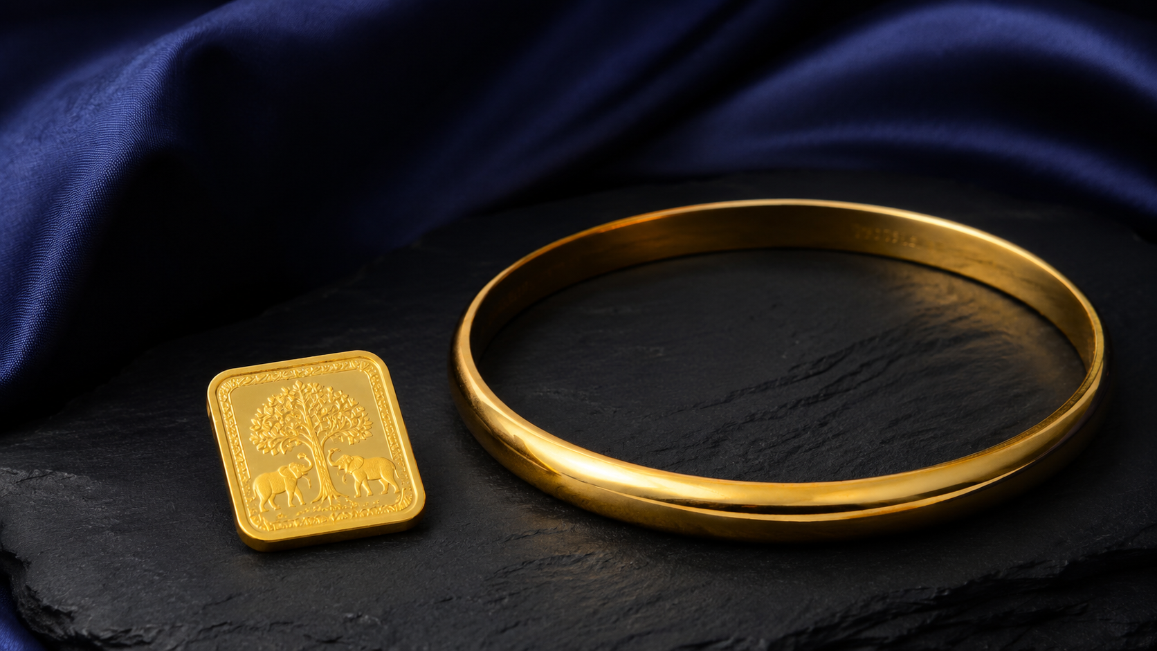 A small minted gold bar beside a plain gold bangle on a dark slate surface